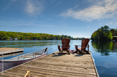 Two Muskoka chairs sitting on a wood dock facing a lake. A canoe is tied to the dock. Life jackets are visible near the chairs.