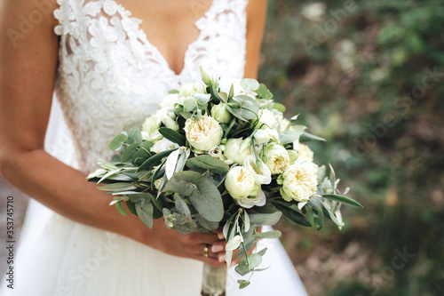 Bride holding white / green wedding bouquet in her hands against white wedding gown. Wedding day concept.