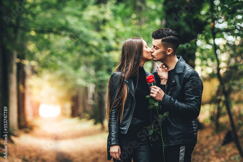Passionate young man and woman kissing in the autumnal park, holding together a red rose.