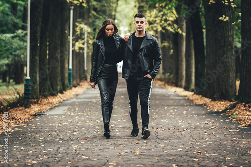 Dramatic shot of couple in love both weard in black leather jackets, holding hands and strolling in autumn park.