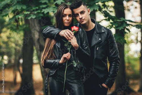 Gorgeous fashionable couple in black posing outside with red rose in hands, looking unemotional and confident.