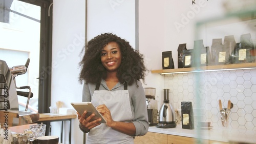 Photography Portrait of female african barista with brown curly hair using the tablet, looking to the camera and smiling in the coffee cafe