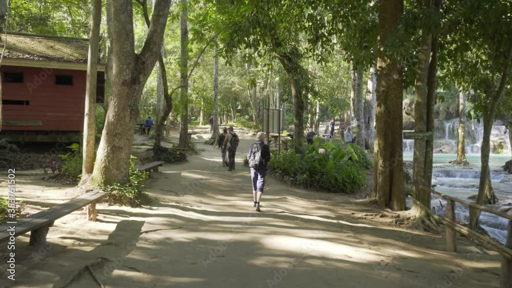 Tourists walking on trail amidst trees by river flowing in forest, famous waterfall in jungle - Luang Phabang, Laos Tourists walking on trail amidst trees by river flowing in forest, famous waterfall in jungle - Luang Phabang, Laos