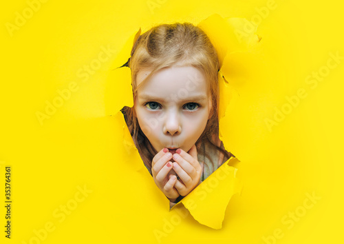 A little funny girl looks out through a hole in torn yellow paper, covers her mouth with her hands and eyes wide in surprise. The concept of wonder, shock, discounts and sales. Background, copy space