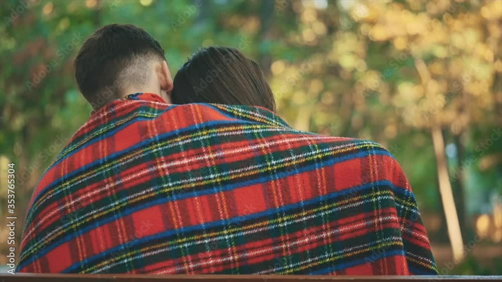 Slowmo. Young pretty couple hugs with cozy blanket, sitting on a bench, on blurred park background. Copy space. Back view. 4K. Slowmo. Young pretty couple hugs with cozy blanket, sitting on a bench, on blurred park background. Copy space. Back view. 4K.