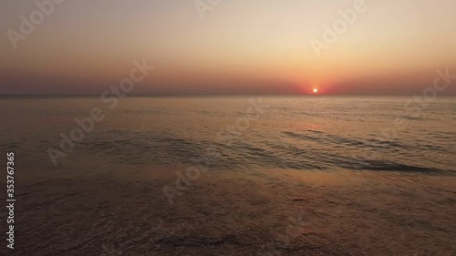 over a sandy beach next to the sea waves slowly rolling in on the shore and the setting sun like a red dot on the horizon