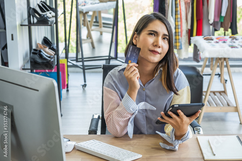 Photography Young Asian woman sitting in front of a computer in a clothing fashion shop looking at tablet holding a credit card with thinking or deciding expression