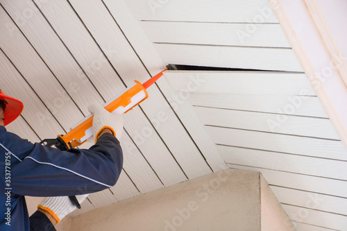 Technician man hand holding and using glue gun, silicone tube or manual caulking gun to repair and fix the wooden cornice with the ceiling at the under roof at home exterior.