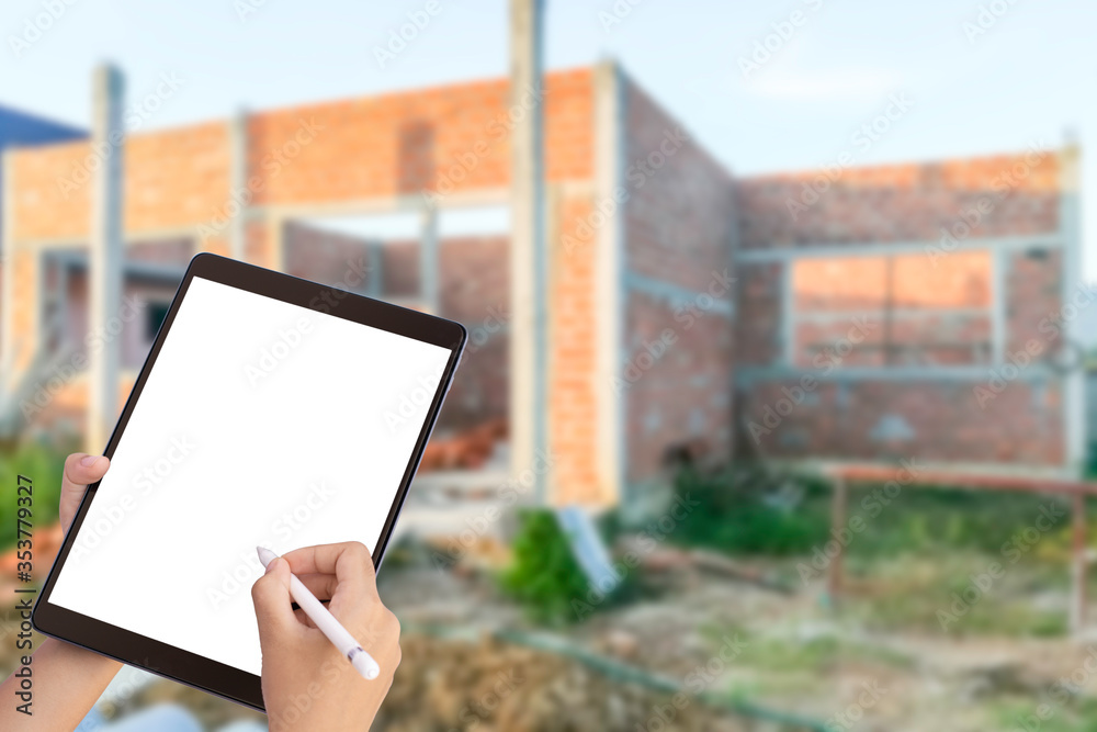 Mockup hand holding and writing on black tablet computer pc with empty screen by pencil with house construction blurred background. Augmented reality (AR ) marketing for architecture concept.