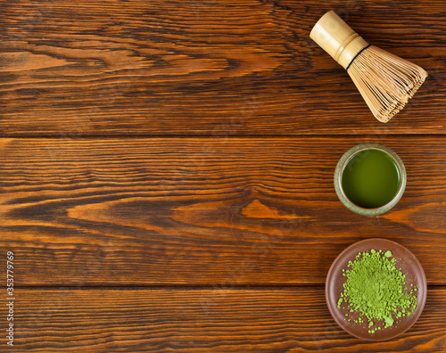 Green matcha tea in a cup with powdered matcha tea in a plate and bamboo matcha tea whisk also know as chasen on a wooden background. Top view. Text space.