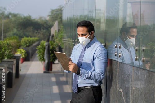 Businessman wearing face mask using his tablet computer outdoors as a mobile office in isolation
