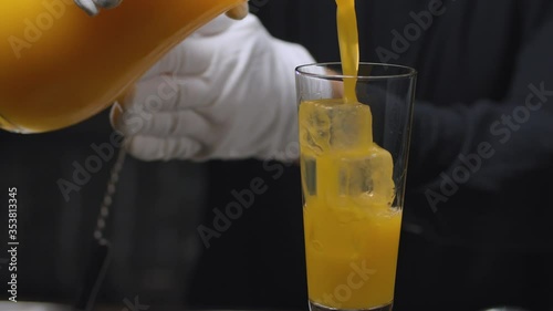 Preparing a Tequila Sunrise Cocktail. Close up bartenders hands pouring orange juice into a glass with ice cubes and garnish with orange and cherry on skewer.