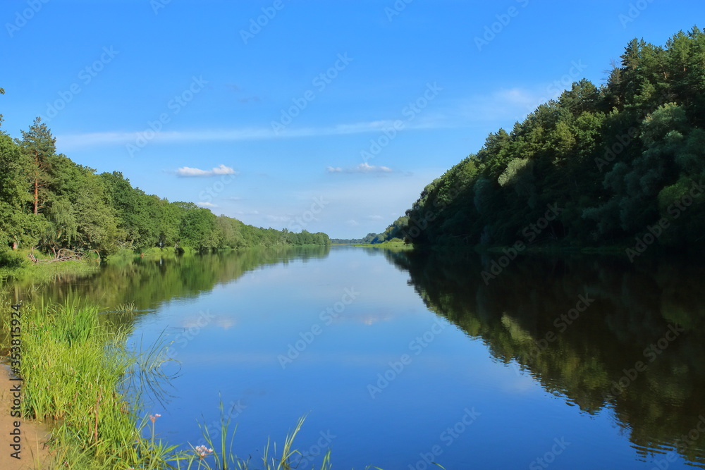 Beautiful view of the wide river from the sandy shore. Morning on the riverbank. The blue sky is reflected in the water. A great place for fishing and recreation. Banks covered with forest.