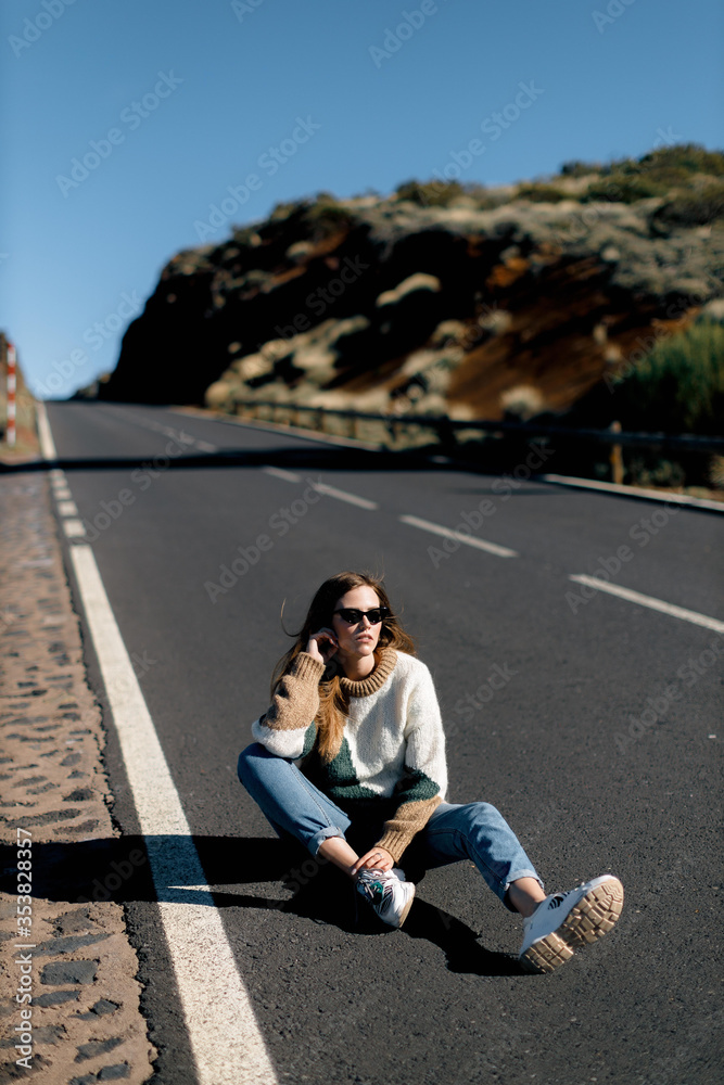 a young girl with long hair in hipster clothing and sunglasses is sitting on an asphalt road