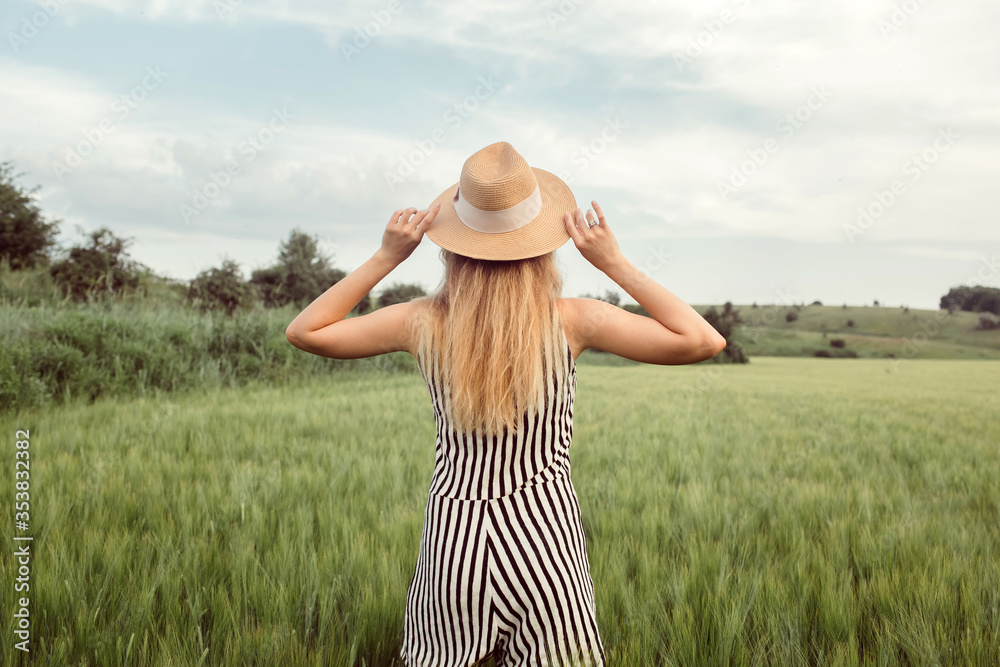 A girl in a straw hat walks on a green field at sunset. Blonde in nature on a summer day. Summer holidays.