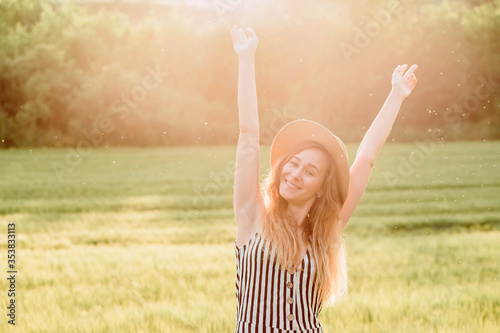 A girl in a straw hat walks on a green field at sunset. Blonde in nature on a summer day. Summer holidays.