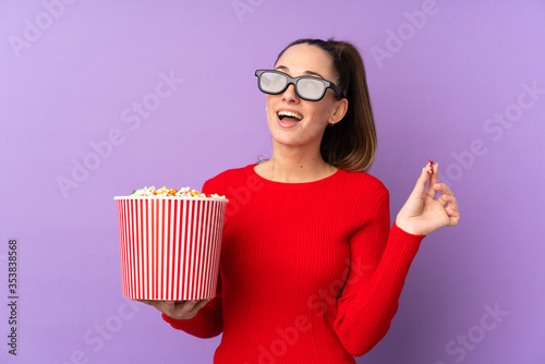 Young brunette woman over isolated purple background with 3d glasses and holding a big bucket of popcorns