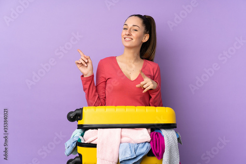 Traveler woman with a suitcase full of clothes over isolated purple background pointing finger to the side