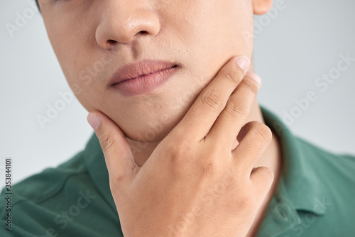 A close up portrait of young thinking man touching his chin with hand and standing against light grey background. Thinking out loud
