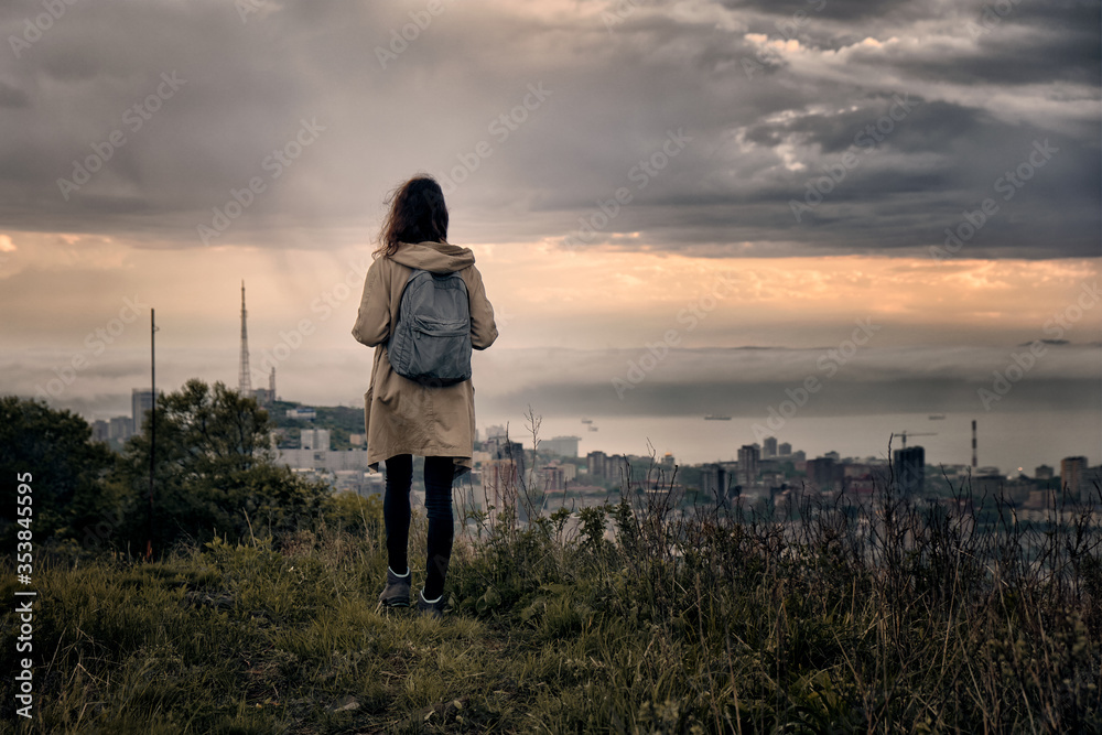rear view of a close up silhouette of a young girl on a hilltop with grass and cloudy sunset sky background and city view. Vacation and outdoor concept.