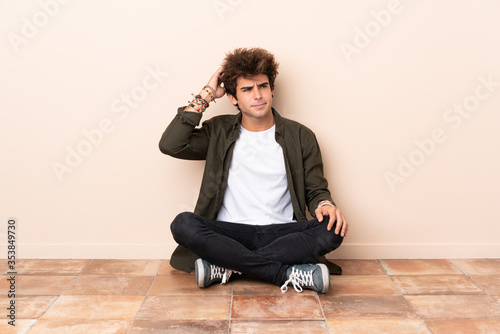 Young caucasian man sitting on the floor having doubts while scratching head