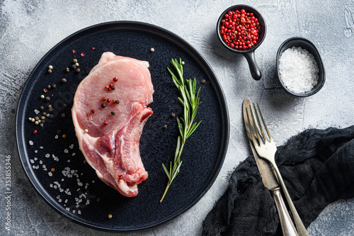 Raw Pork Loin chops  in a black round plate on a grey textured  stone background with rosemary garlic peppercorns ingredients for grilltop view photo