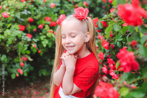 Outdoor portrait of a pretty girl of 6-7 years old against the background of a flowering red rose. A girl with long blond hair in a beautiful red dress. Little girl makes a wish closing her eyes