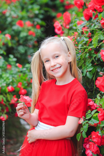 Outdoor portrait of a cute girl of 6-7 years old against the background of a blooming red rose. Girl with long blond hair in a beautiful red dress. Spring flowering roses in the city