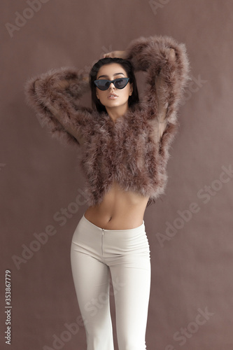 Young elegant girl posing in fluffy pink cape with artificial fur and beige flare trousers on brown background