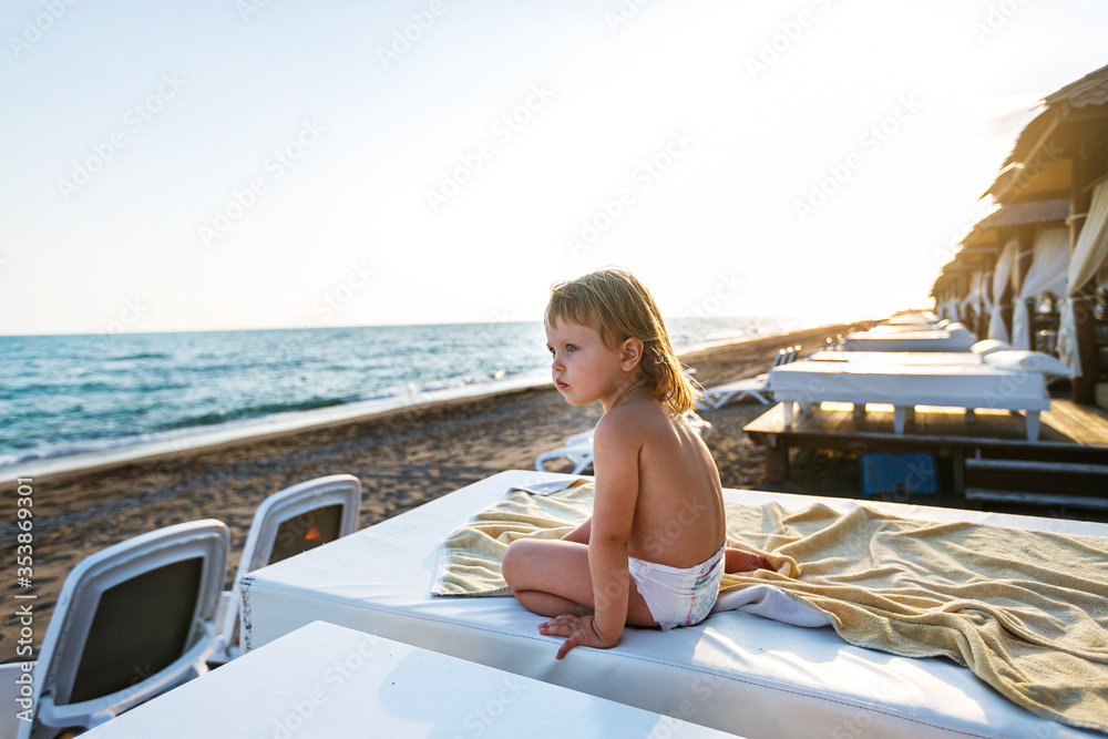 A little happy girl in a diaper on the sea in a VIP Bungalow sitting looking out at the sea at sunset. Family holidays with children at the sea