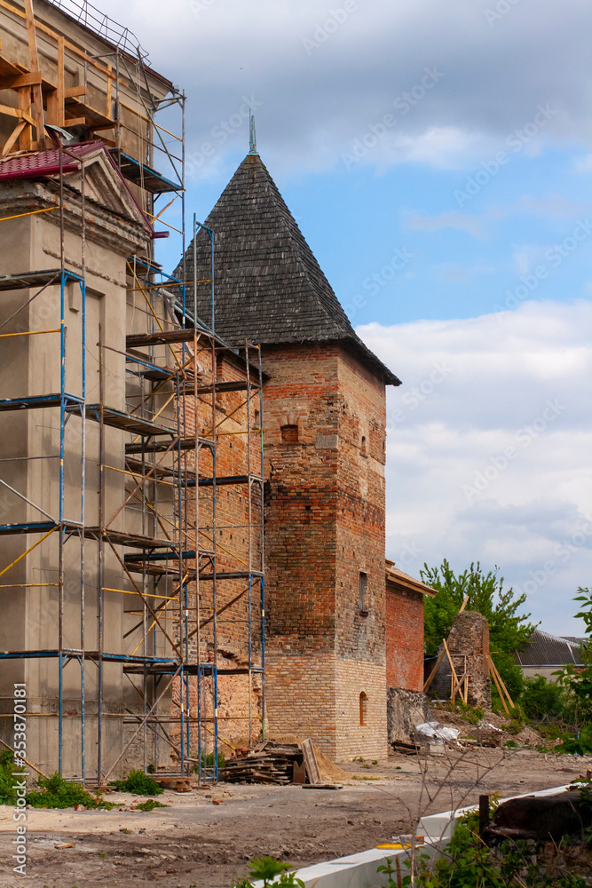 Scaffolding on the wall of an old castle. Restoration of architecture. Repair work of the monastery and castle wall. Old tower of a medieval castle with a wooden roof. Vertical photograph.