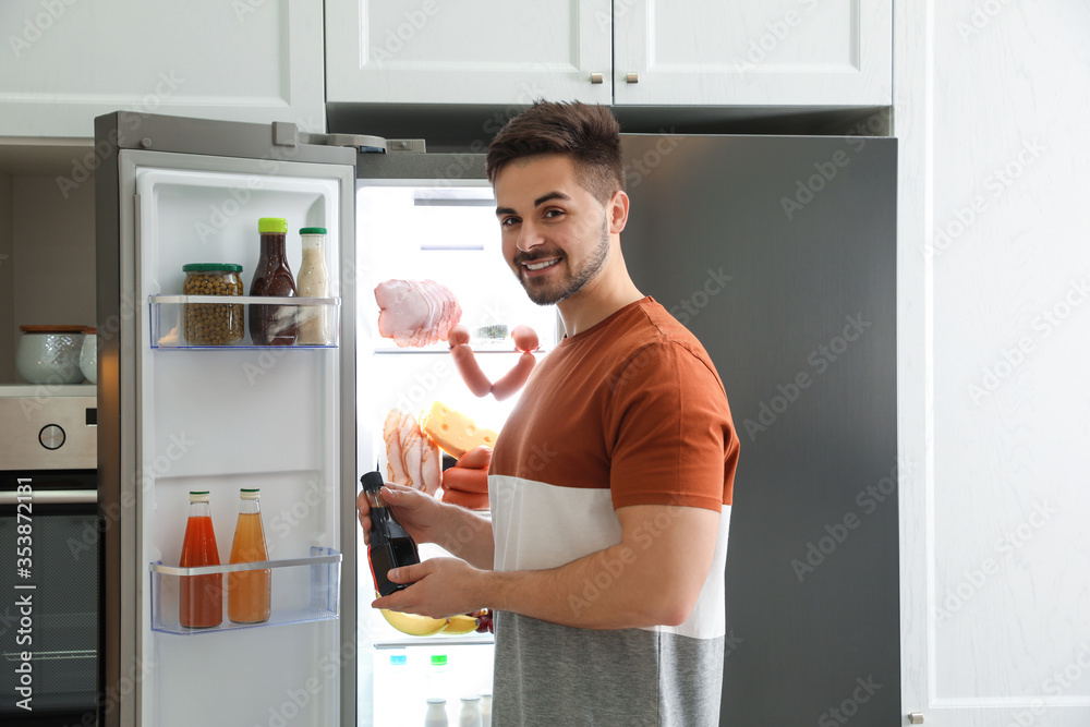 Young man with bottle of sauce near open refrigerator indoors Young man with bottle of sauce near open refrigerator indoors
