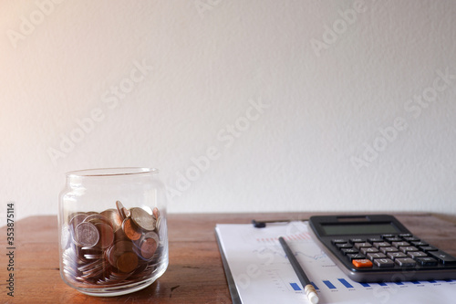 The cropped view of the glass jar contains the coins withe calculator and business paper on the wood desk with white natural blackground for texing the message, information,and background. photo