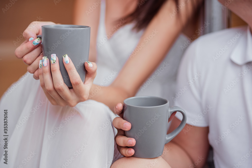 Two lovers drink hot drinks from the gray cups that they hold in their hands. A beautiful girl and a young man are sitting on the doorstep of their trailer truck and enjoying outdoor recreation. Two lovers drink hot drinks from the gray cups that they hold in their hands. A beautiful girl and a young man are sitting on the doorstep of their trailer truck and enjoying outdoor recreation.