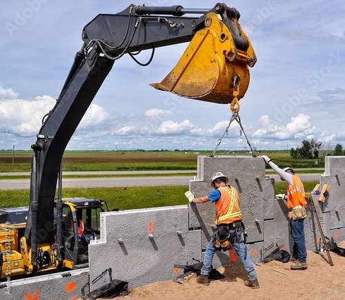 Mechanically Stabilized Earth (MSE) wall construction with an excavator lifting a concrete wall panel white two workers guide the panel into place and onto nylon pins to ensure proper installation.