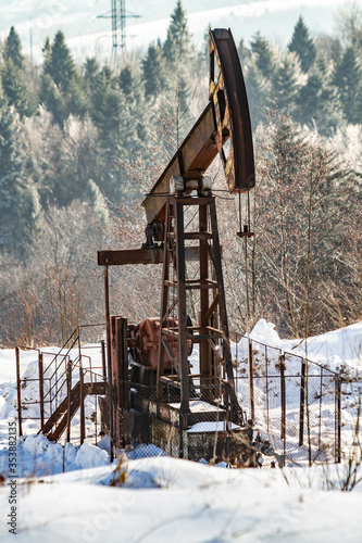 An old oil rig pumps oil against the backdrop of snowy trees and mountains on a sunny day. Vertical orientation.
