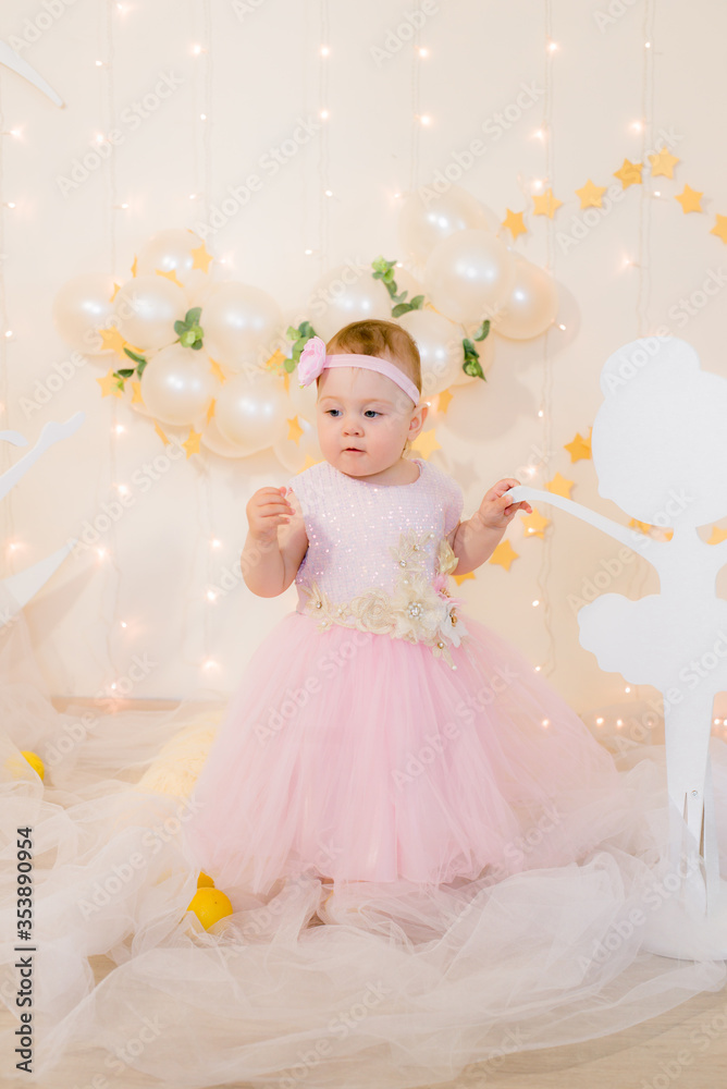 Fototapeta premium Cute little girl in a pink princess dress with a flower on her hair plays with wooden ballerinas among the golden stars on the background of a large white wooden moon and balloons. Children's decor. 