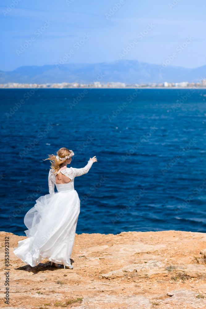 Obraz premium Bride with wreath in white wedding dress on the rock beach against beautifull blue sea background. Blond bride pointing at sea on the beach. Concept of wedding and bridal morning. wedding Mallorca,