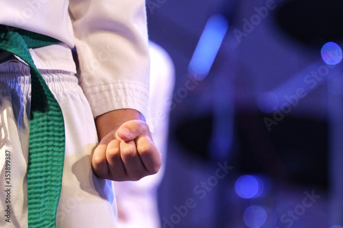 Taekwondo kid player make fist of left hand at beside the hip, wear Taekwondo white uniform and green belt. Background is blur fist of other player, image blue tone from blue light decoration of stage