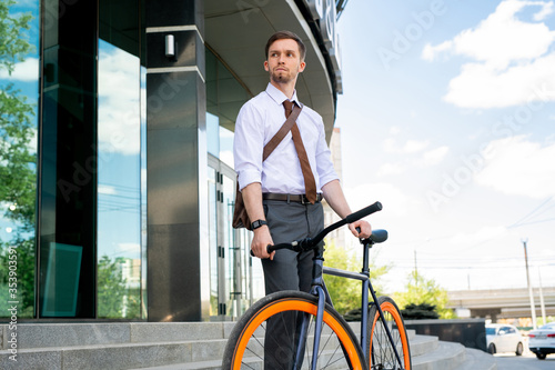 Young serious businessman going to go by bicycle while standing outdoors