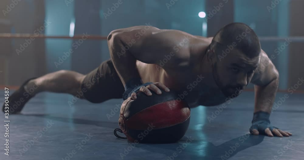 Close up of male Caucasian boxer warming up in dark ring. Strong man doing difficult push ups in darkness. Guy kickboxer pushing up with difficulties on one arm before training.