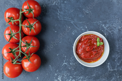 Tomato sauce-ketchup in a bowl with spices, herbs and fresh cherry tomatoes on a branch, on a dark graphite background. The view from the top.