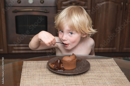 .little boy with blond hair sits at a table in the kitchen and eats with a spoon a chocolate dessert and look at the camera