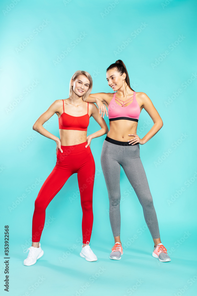 Ready for workout. Full length of two young and attractive sporty girls in sportswear looking at camera and smiling while standing against blue background. Sport and fitness Ready for workout. Full length of two young and attractive sporty girls in sportswear looking at camera and smiling while standing against blue background. Sport and fitness