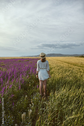 Escape. Young woman in hat posing in a wheat field with purple Consolida flowers on a summer day