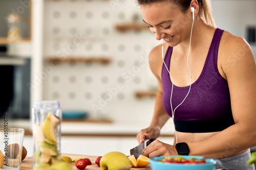 Happy sportswoman slicing fruit while preparing smoothie in the kitchen.
