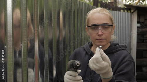 A woman in work clothes and gloves holds and examines an electric screwdriver