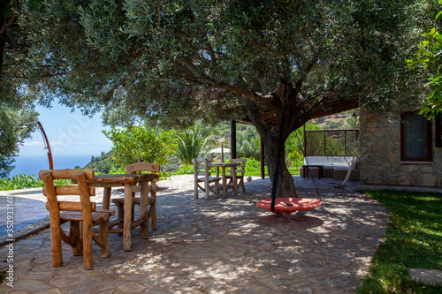 Wooden chairs and tables, red swing in a beautiful garden with beautiful trees and sea view.