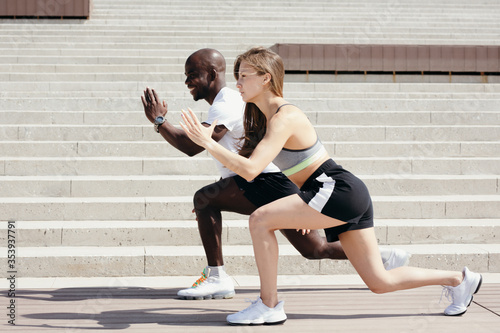 African American coach trains the blonde athlete in the outdoors. Exercise a lunge to stretch the quadriceps.