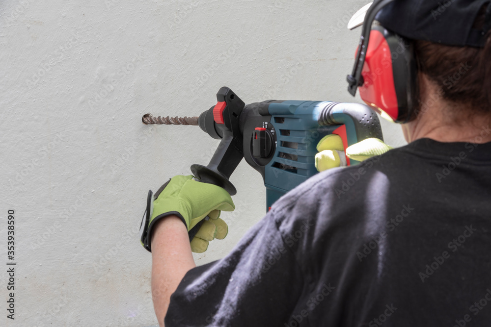 Fototapeta premium Mujer usando un martillo rotativo en el trabajo, abriendo un agujero en una pared blanca, usando guantes y protectores auditivos